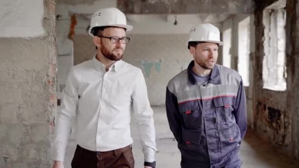 Hauptmeister und Inspektor auf der Baustelle. der Bauunternehmer trägt eine spezielle Uniform und zeigt mit einem Helm auf dem Kopf dem Inspektor die geleistete Arbeit. — Stockvideo