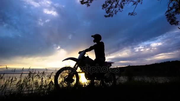 Silueta de un conductor de transporte extremo que se sienta en una motocicleta, un vehículo se para cerca de un río sobre un fondo de puesta de sol — Vídeo de stock