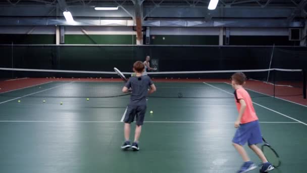 Тренер-женщина в спортивном костюме учит играть в теннис у двух мальчиков среднего возраста, парни прыгают мячами на теннисном корте — стоковое видео