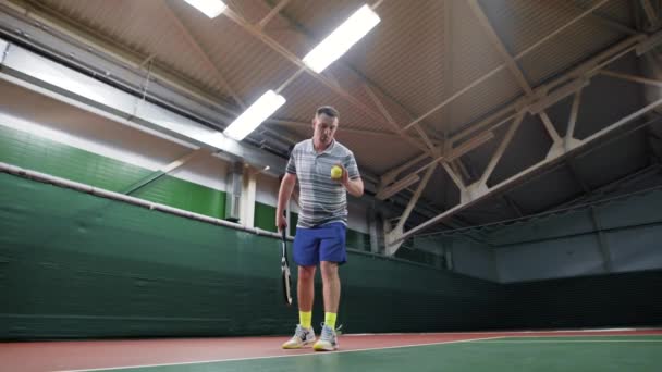 Een volwassen mannetje in sporten kleren serveert een serve met een racket en een tennisbal, de persoon is in een gesloten stadion van een sportcomplex — Stockvideo