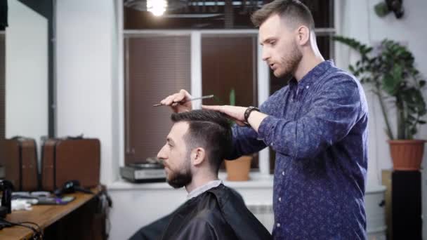 Um cabeleireiro elegante penteia suavemente o cabelo de seus clientes, a fim de criar um penteado elegante em um salão de beleza para o visitante com uma barba — Vídeo de Stock