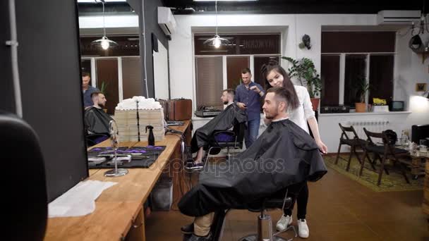 En una peluquería de moda hay un corte de pelo de varios visitantes, los hombres están esperando el final de sus cortes de pelo y peinado con la ayuda de gel y otros estilos — Vídeo de stock