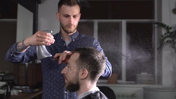 El peluquero salpica el agua del atomizador en la cabeza del visitante de la peluquería que está esperando a que termine el corte de pelo. — Vídeos de Stock