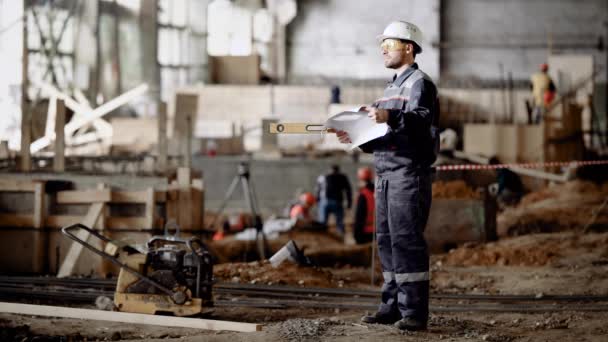Trabalhador masculino no local. Vista lateral do homem de uniforme em pé com o plano do projeto em mãos no fundo do site . — Vídeo de Stock