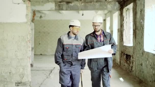 Deux travailleurs collaborent au plan du projet. Deux hommes en uniforme et chapeaux durs discutant du nouveau projet et travaillant avec le plan directeur . — Video
