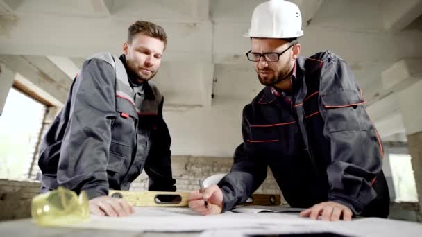 Подрядчик и рабочий, работающие за столом. Двое мужчин в форме работают с планом нового проекта, стоя за столом на фоне площадки . — стоковое видео