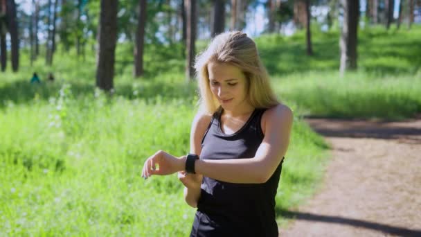 Женщина в спортивной одежде смотрит на часы во время пробежки в зеленом лесу — стоковое видео