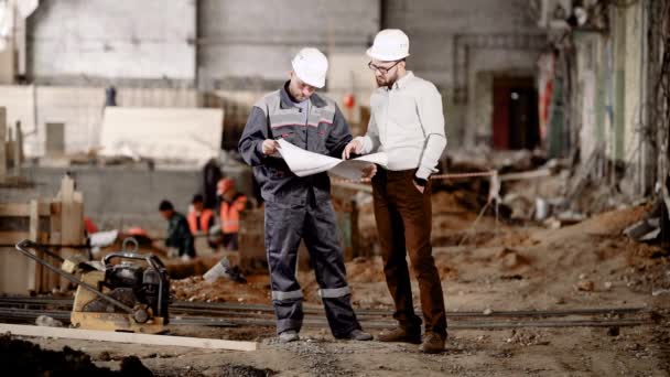 Dos hombres en cascos de seguridad de pie en el área de construcción y mirando el esquema de la futura construcción. Capataz apuntando a dibujar y explicando a los trabajadores lo más destacado del ensamblaje actual . — Vídeos de Stock