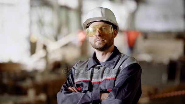 Närbild av arbetande mannen i uniform, skyddande hardhat och glasögon står i byggande området och tittar mot kameran. Ung man arbetar i byggplats, leende och gestikulerande tummen upp. — Stockvideo