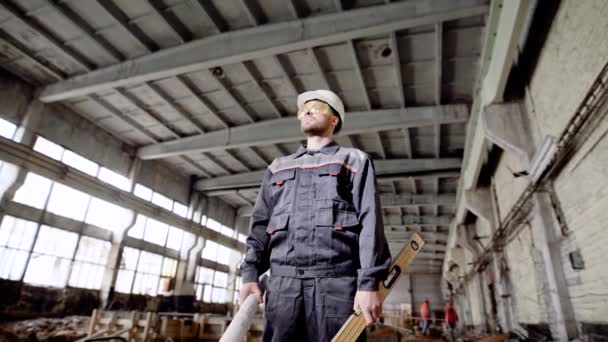 Junger Polier im Baubetrieb, im Hintergrund Schweißarbeiten. Arbeiter hält Bauplan und Wasserwaage in der Hand, überprüft den Bauablauf und blickt über den Arbeitsbereich. — Stockvideo