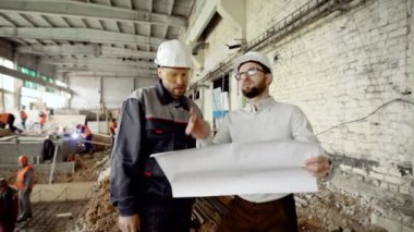 İki erkek Koruyucu baretler, inşaat alanı içinde arka planda kaynak işçiler ile birlikte çalışıyoruz. Foreman ve mimar site oluşturma ve yapısal çizim holding ayakta.