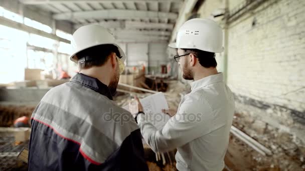 Achteraanzicht van mannen in helmen en brillen permanent samen in Bouwoppervlakte met mensen op de achtergrond. Architect en werknemer Structuurplan kijken en bespreken van de details voor ontwikkeling. — Stockvideo