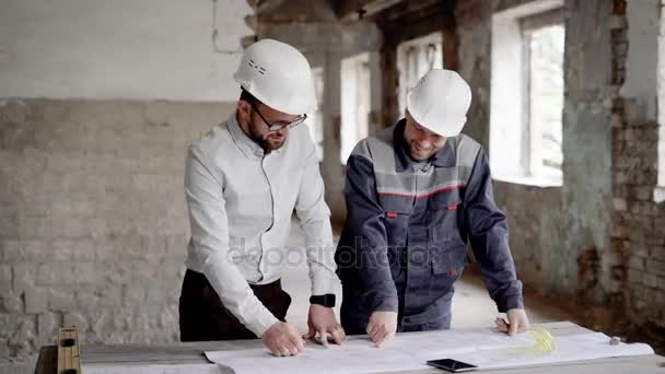 建築家と構造エンジニアは、テーブルの上、計画と設計図を議論しています。建物面積互いに話しているし、スキームを指して hardhats で 2 人の男性が立っています。. — ストック動画