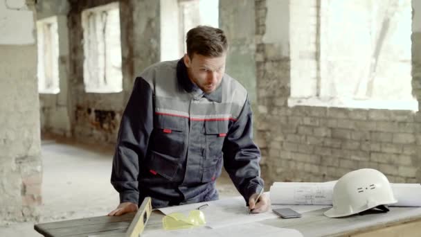 Der junge Polier steht auf der Baustelle und zeichnet den Bauplan. Bauingenieur arbeitet mit Planungsunterlagen. Architekt erstellt Entwicklungsplan am Schreibtisch. — Stockvideo