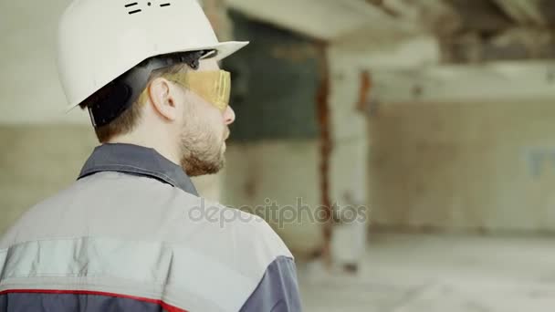 Back view of inspector dressed in uniform, protective hardhat and eyeglasses walking on the building site and checking industrial project. Professional surveyor is inspecting construction area. — Stock Video