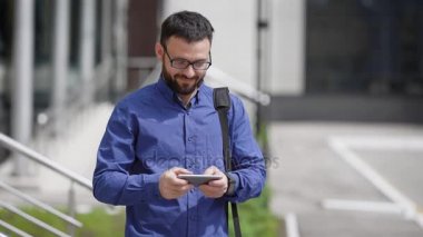 Yakışıklı sakallı adam mavi gömlek giymiş ve kahverengi pantolon açık havada duran ve onun smartphone kullanarak ileti yazarak. Genç işadamı arkadaşlarım sokakta güneşli bir günde bekliyor.