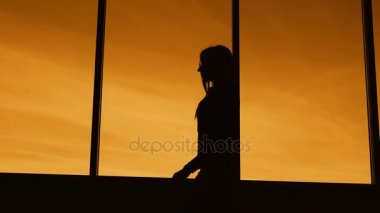 Karanlık siluet alacakaranlık panoramik kat tavan penceresi boyunca yürürken profil içinde uzun saçlı bir ince kadın. Anahat arka planda turuncu günbatımı gökyüzü karşı kadın vücudunun.