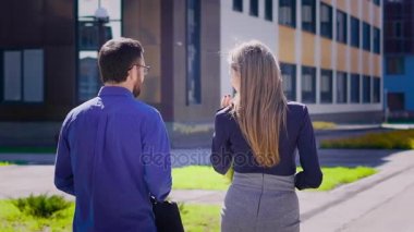 İşadamı ve birlikte yürüyüş iş kadını arkadan görünüm. Kadın meslektaşı ile bina boyunca bir yürüyüş alarak tişörtlü yakışıklı adam. Zarif Bayan bluz ve etek onun arkadaşı için dinleme.