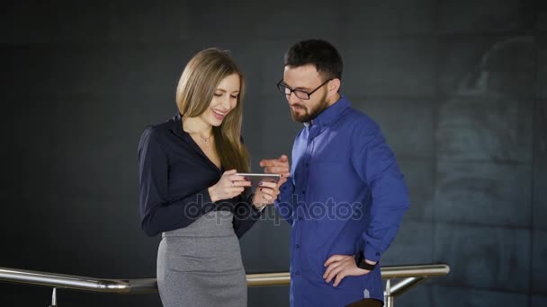 Two colleagues are chatting in the office and sharing impressions about their job. Man dressed in blue shirt is listening to woman in smart suit with smartphone in her hands. — Stock Video