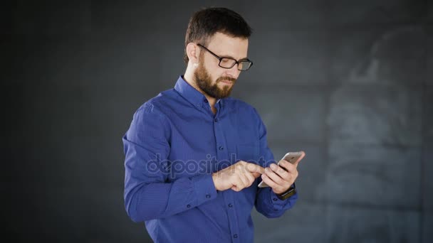 Close up de homem barbudo bonito em óculos elegantes, camisa azul em pé no escritório e trabalhando usando smartphone. Jovem empresário com relógio de pulso preto digitando mensagens no dispositivo digital . — Vídeo de Stock