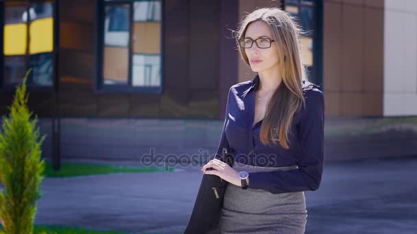 Giovane bella signora vestita con camicetta blu e gonna grigia sta avendo una passeggiata all'aperto lungo il moderno edificio per uffici con borsa attraverso il suo shouder. La donna d'affari sta andando alla conferenza . — Video Stock