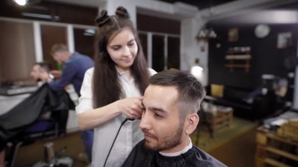 Mulher barbeiro aparar o cabelo em seu cliente com cortador de cabelo. Cena da barbearia — Vídeo de Stock
