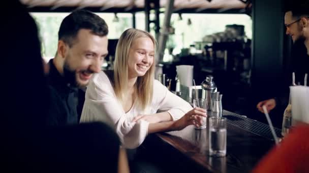 Молодые друзья, которые сидят на барных стульях рядом со стойкой бора общаться друг с другом, красивая блондинка рассказывает смешную историю и улыбается — стоковое видео