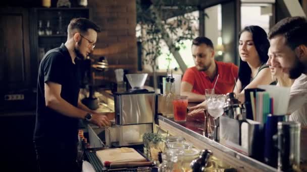 O jovem barman adiciona gelo ao coquetel para esfriá-lo uma empresa de amigos esperam suas bebidas, amigos querem se divertir no bar — Vídeo de Stock