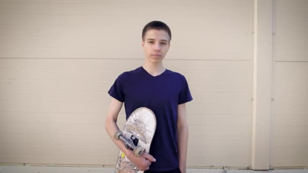 Imagen de un adolescente guapo parado en la calle y sosteniendo el monopatín en sus manos. Chico pasar tiempo libre en el parque de skate demostrando un estilo de vida saludable y mirando a la cámara . — Vídeo de stock