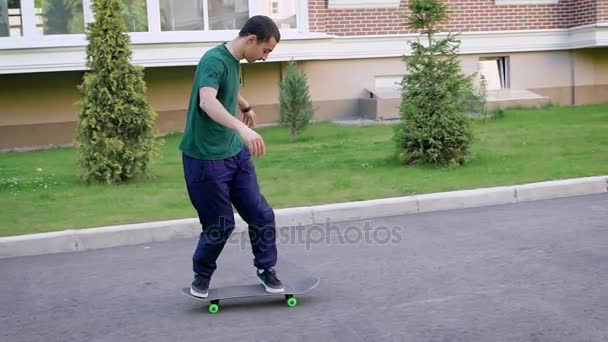 Närbild av unga skateboardåkare klädd i casual grön tröja, blå byxor och sneakers ridning på plan yta i sommardag. Mannen skridskor längs vägen hoppar upp gör tricks ombord. — Stockvideo