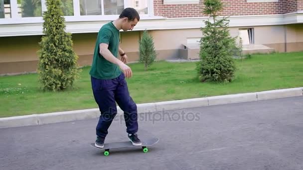 Close up van jonge skateboarder gekleed in casual groen shirt, blauwe broek en sportschoenen rijden op vlakke ondergrond in zomerdag. Man schaatsen langs de weg is springen omhoog doend trucs aan boord. — Stockvideo