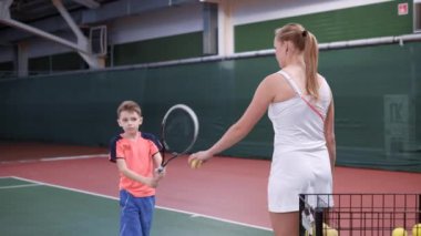 Profesyonel ekipman ile kapalı saraydaki resim dersin. Çocuk öğrenme sürecinde doğru bir raket kullanarak. Çocuk beyaz kıyafet onu sarı topları veren kadın Koç Tenis oynarken.