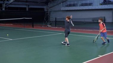Kapalı sarayda oğulları Tenis oynarken annesi. Rekreasyon alanı içinde profesyonel spor oyun oynayan çocuklar öğretim koç. Genç sporcuların dönen ve sarı topları raketle hizmet öğrenme.