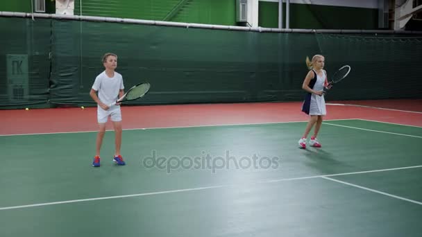 Twee gelukkige kinderen dienen en terugkeer ballen opleiding vaardigheden op Hof met rackets. Mooie jongen en meisje sport spel in recreatiegebied. Toekomstige professionele spelers die van tennis les. — Stockvideo