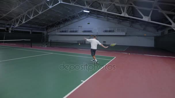 Крупный план красивого профессионального теннисиста в белой рубашке, серые шорты и обувь тренировки на крытом корте с оборудованием. Мужчина подает и возвращает мячи с рейнджерами в зоне отдыха . — стоковое видео