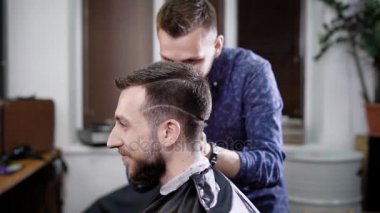 Yan görünümü şık gülümseyen bir kafa ile siyah sabahlığı kaplı berber içinde oturan adam sakallı. Berber boxer saç kesme makinesi ile yapma müşteri arkasında, rahat kıyafetler içinde ve tarak.