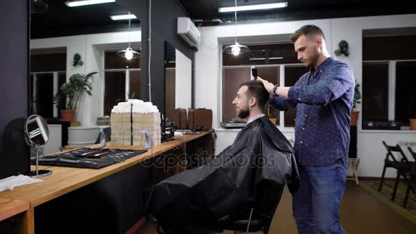 Joven peluquero con ropa casual de pie y peinando al cliente antes de cortarse el pelo. Hombre sonriente sentado en la silla cubierta de peignoir negro y mirando el espejo frente a la barbería . — Vídeos de Stock