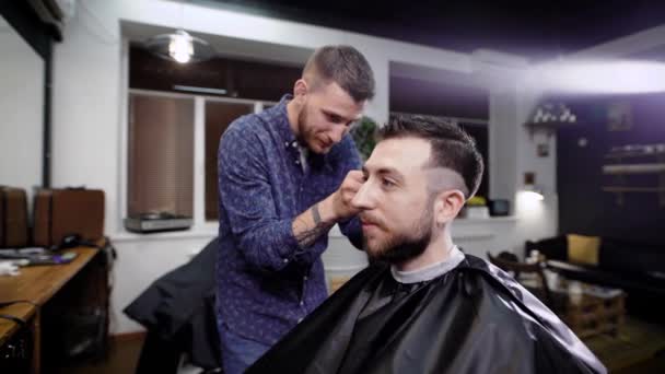 Primer plano de hombre barbudo sonriente elegante sentado cubierto con peignoir negro en el barbeshop. Peluquero vestido con ropa casual de pie detrás del cliente y haciendo corte de pelo boxeador con clipper y peine . — Vídeos de Stock