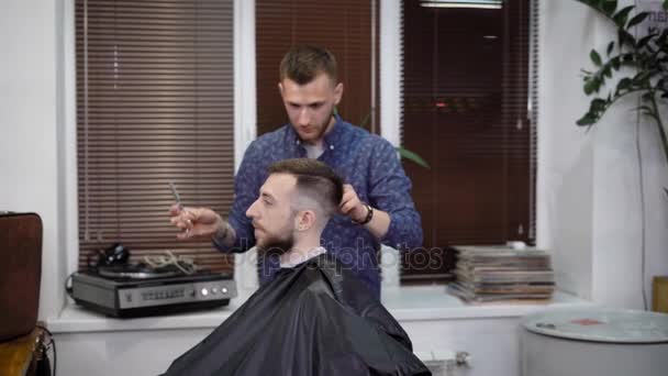 Homme coiffeur travaillant avec le client recouvert d'une cape de coupe façonnant parfaitement sa coupe de cheveux avec peigne et ciseaux. Femme coiffeuse utilisant tondeuse électrique pour faire coiffure moderne de bel homme . — Video