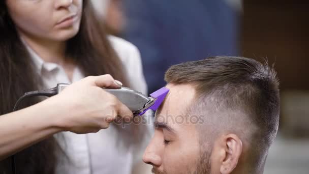 Primo piano di parrucchiere femminile professionale vestito con abiti casual sta servendo cliente in barbiere utilizzando trimmer moderno. Giovane uomo sta ottenendo taglio di capelli alla moda seduto sulla sedia chiudendo gli occhi . — Video Stock