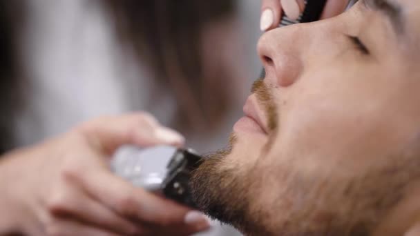 Крупным планом молодой бородатый мужчина стрижется на лице у парикмахера в стильной парикмахерской. Изображение женских рук, использующих триммер, получающих идеальную форму для бороды и усов красивого хипстера . — стоковое видео