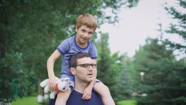 Padre llevando a su hijo de vuelta al parque. Retrato de padre feliz dando paseo hijo sobre sus hombros y mirando hacia arriba. Lindo chico con papá y juguete jugando al aire libre . — Vídeos de Stock