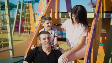 Mutlu bir aile eğlence birlikte oyun alanında yaz gün vakit. Mather, baba ve oğul kayma merdivenlerde oturan kucaklayan ve gülerek kurulu. Anne ve çocuk dışarıda eğleniyor.