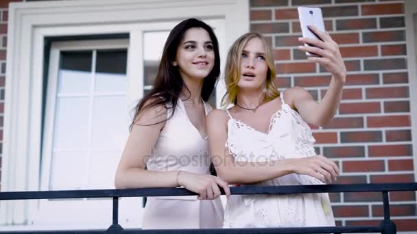 Two wonderful women in white dresses are taking selfie on a smartphone on background of brick wall and window — Stock Video