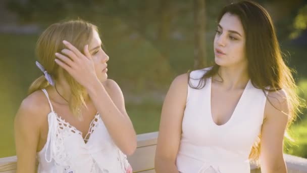 Två goda vänner har ett hjärta till hjärta samtal ute på en parkbänk. Flickor som bär vita klänningar diskutera saker — Stockvideo