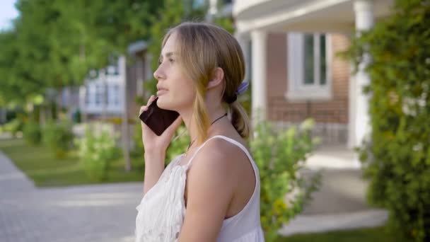 Улыбающаяся милая молодая женщина в белом кружевном платье ходит и разговаривает по мобильному телефону со своей подругой летом — стоковое видео