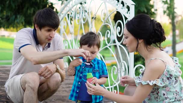 Los padres jóvenes están enseñando a su hijo a soplar burbujas, mamá sostiene un frasco de líquido, papá tiene miedo de soplar, bebé le gusta un nuevo juego — Vídeos de Stock