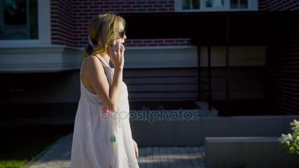 Fiatal üzletasszony sétál egy utca mentén homlokzatok a házak, és beszél telefonon, süt a nap — Stock videók