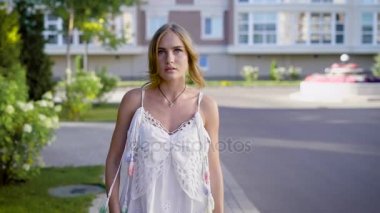 Şık genç sarışın beyaz giyen elbise geziniyor bir sokakta güneşli bir günde, kameraya bakıyor