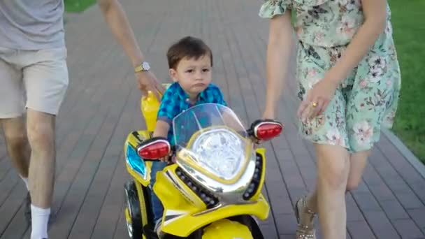 Schöne Familie geht im Park spazieren, während ihr kleiner süßer Sohn entzückendes Motorrad fährt — Stockvideo